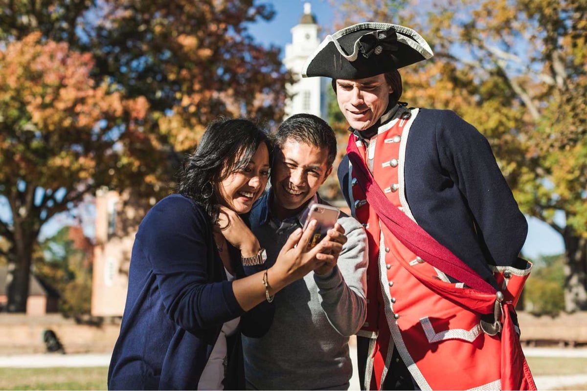 a Revolutionary War era soldier in a tri-corner hat and a modern couple look at the screen of a smartphone