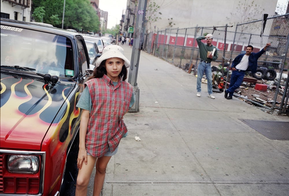 a young girl in a white hat poses next to a car with flames painted on the hood