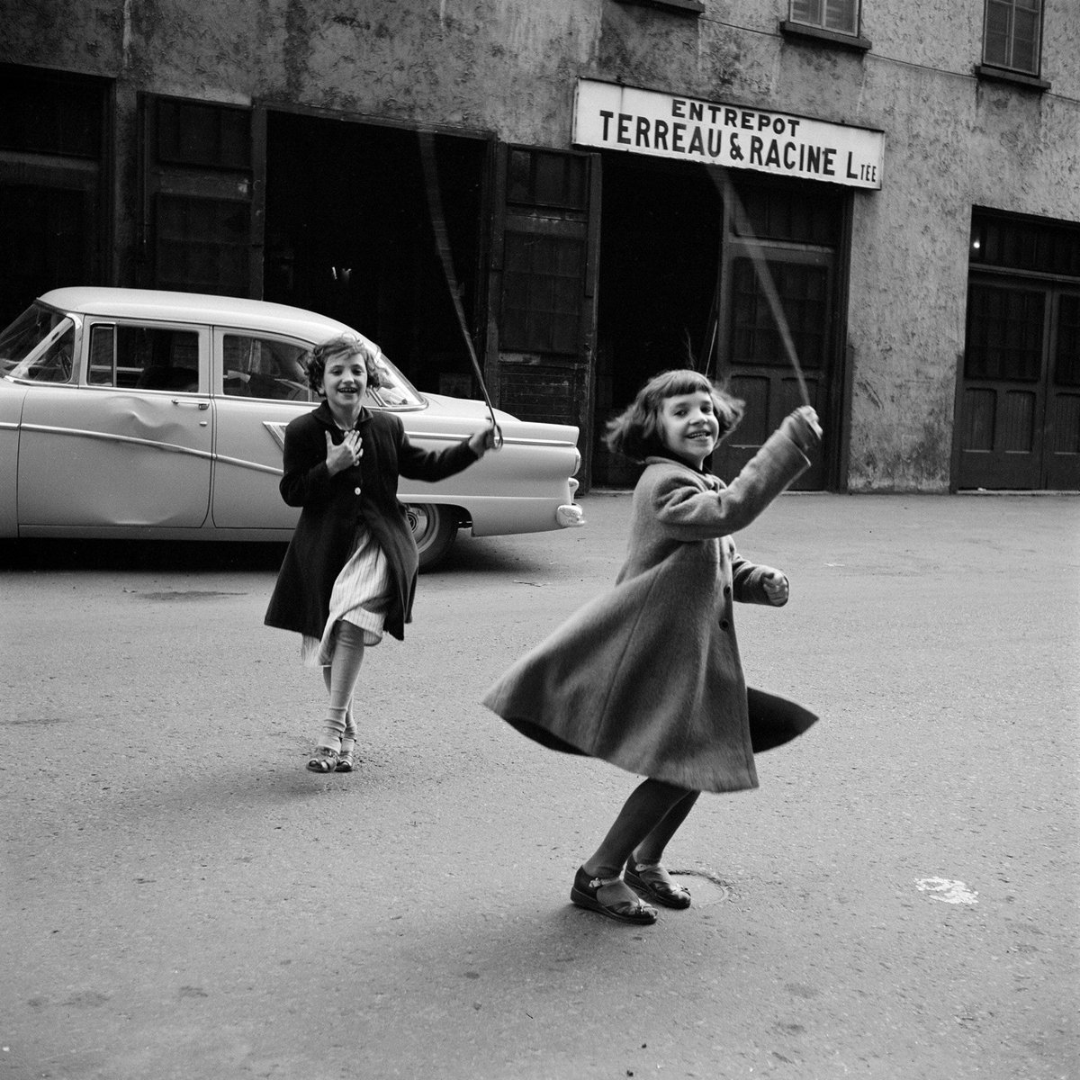 black & white photo of two girls playing on the street in front of a car