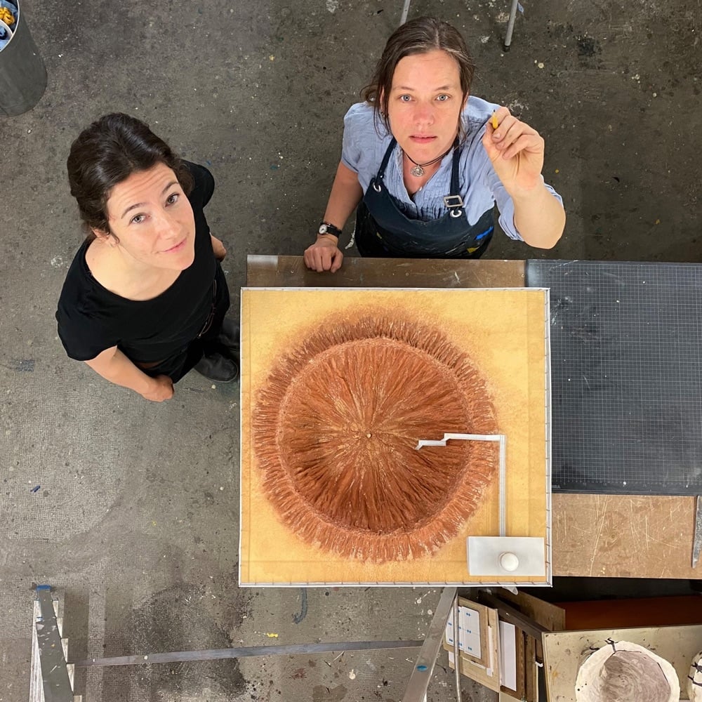 a model of an asteroid impact crater next to two model makers